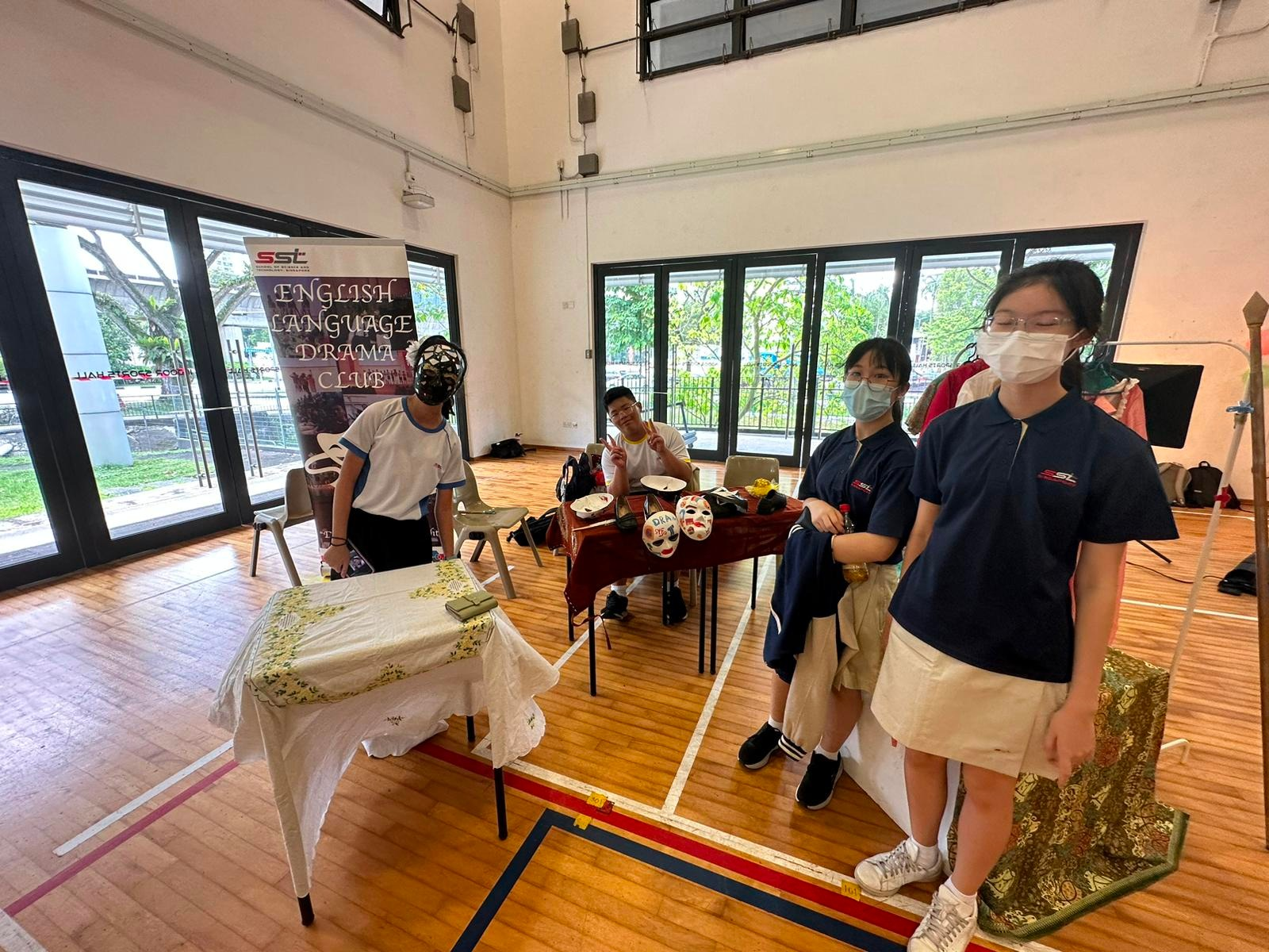 Students in school uniforms stand near a table with masks at an English Language Drama Club booth in a hall.